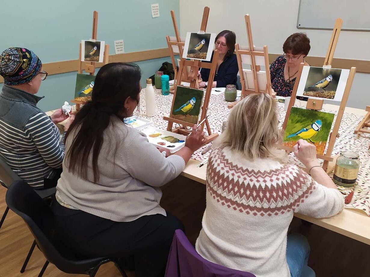 People seated around a table painting small blue tit birds on canvases mounted on easels in a classroom setting