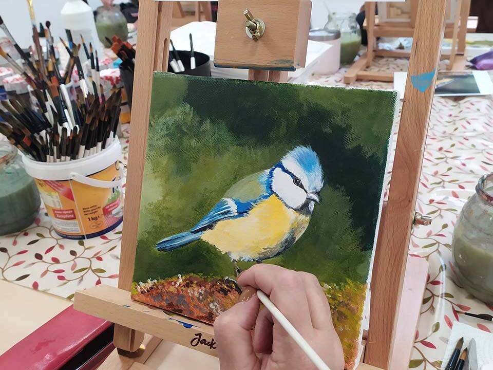 Close-up of a hand painting a blue tit on a green background at an art class easel, with brushes nearby.