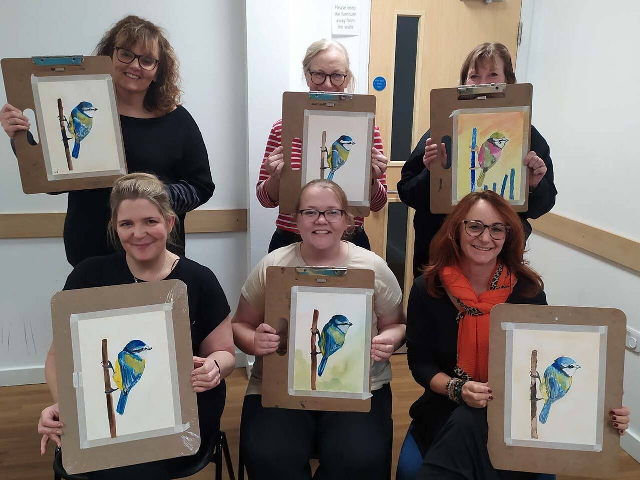 Six women pose in a room, each holding a clipboard with a painting of a blue bird perched on a branch (watercolor).