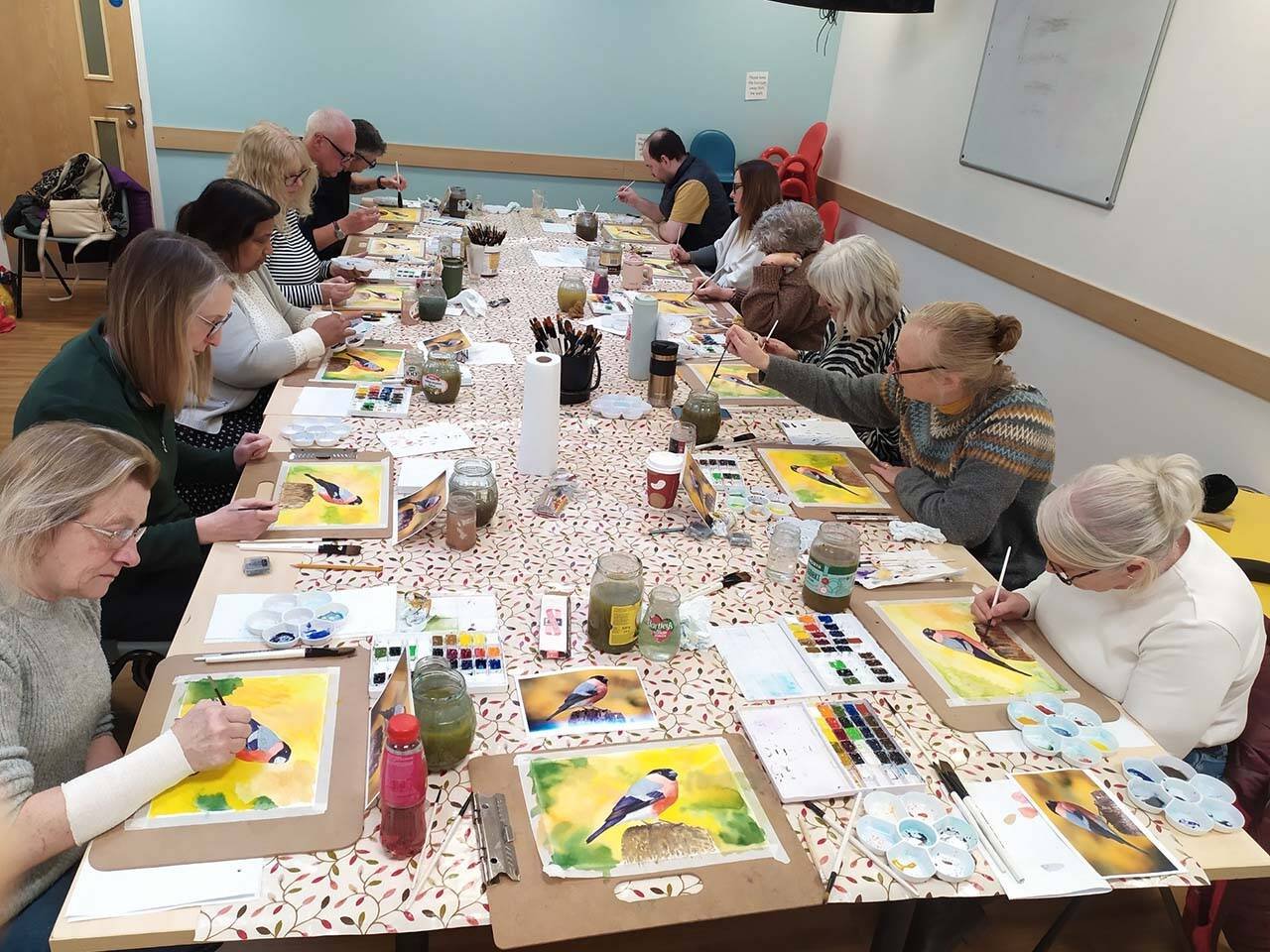Group of adults around a long table painting colorful bird watercolors in a class setting. They work with brushes, paints, and jars of water on a patterned tablecloth.