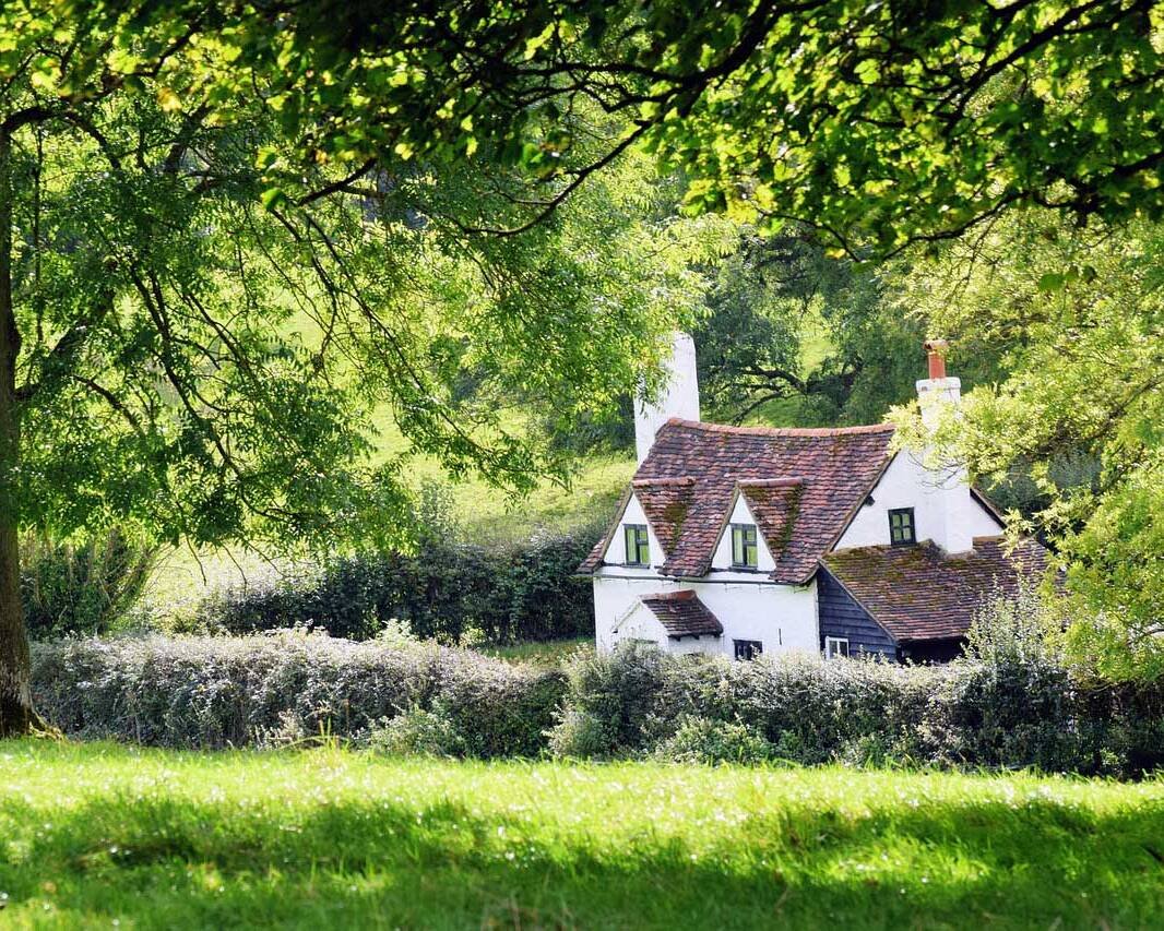 White cottage with a red-tiled roof nestled in a lush green meadow framed by trees and foliage.