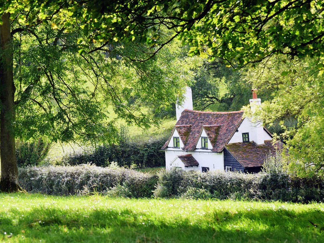 White cottage with a red-tiled roof nestled in a lush green meadow framed by trees and foliage.