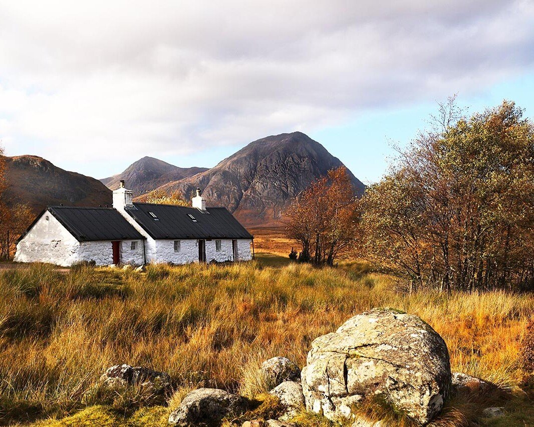 White stone cottages with black roofs sit in a golden autumn field, mountains in the distance under a blue sky.