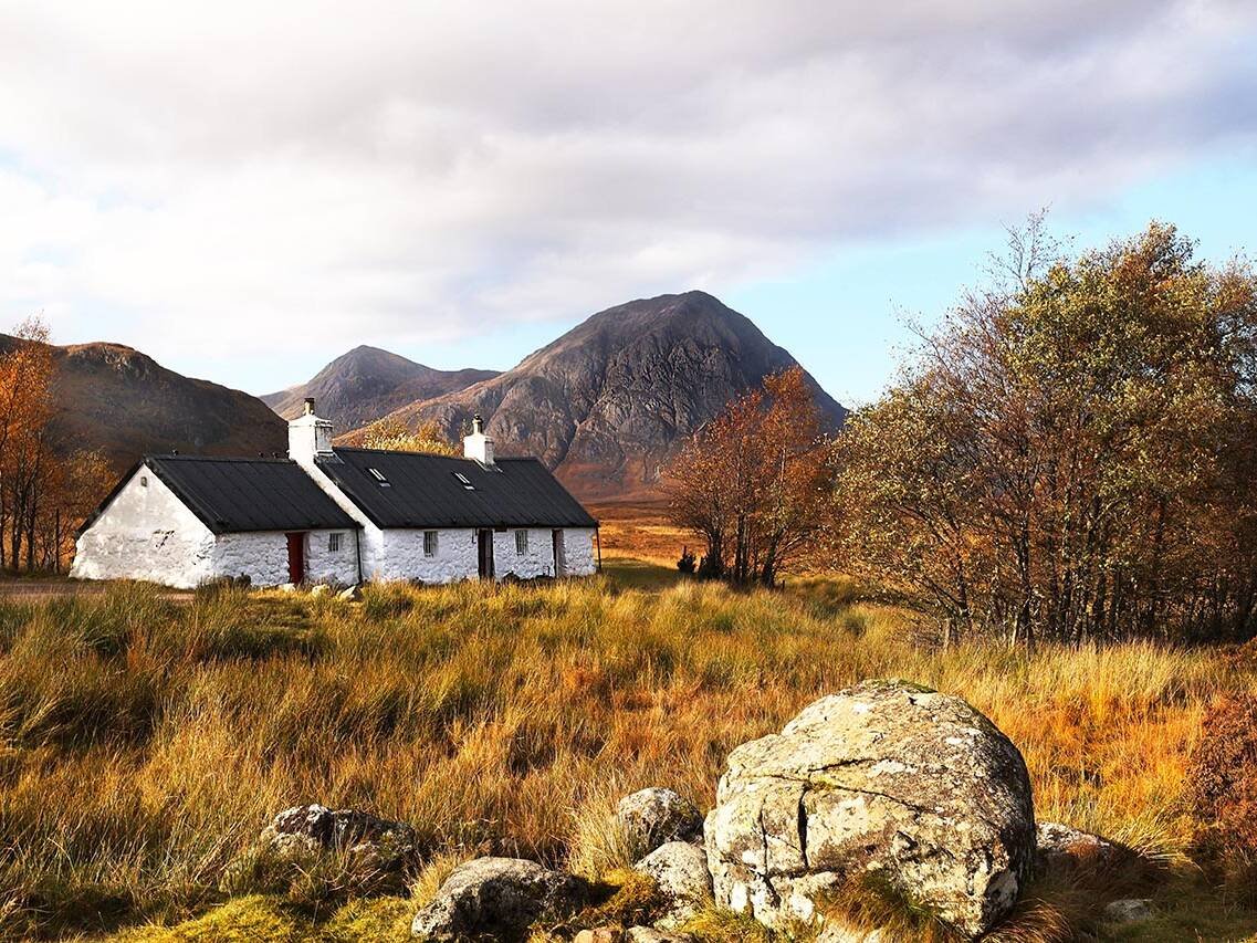 White stone cottages with black roofs sit in a golden autumn field, mountains in the distance under a blue sky.