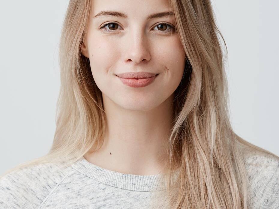 Head-and-shoulders portrait of a smiling young woman with long blonde hair in a light gray sweater against a neutral background
